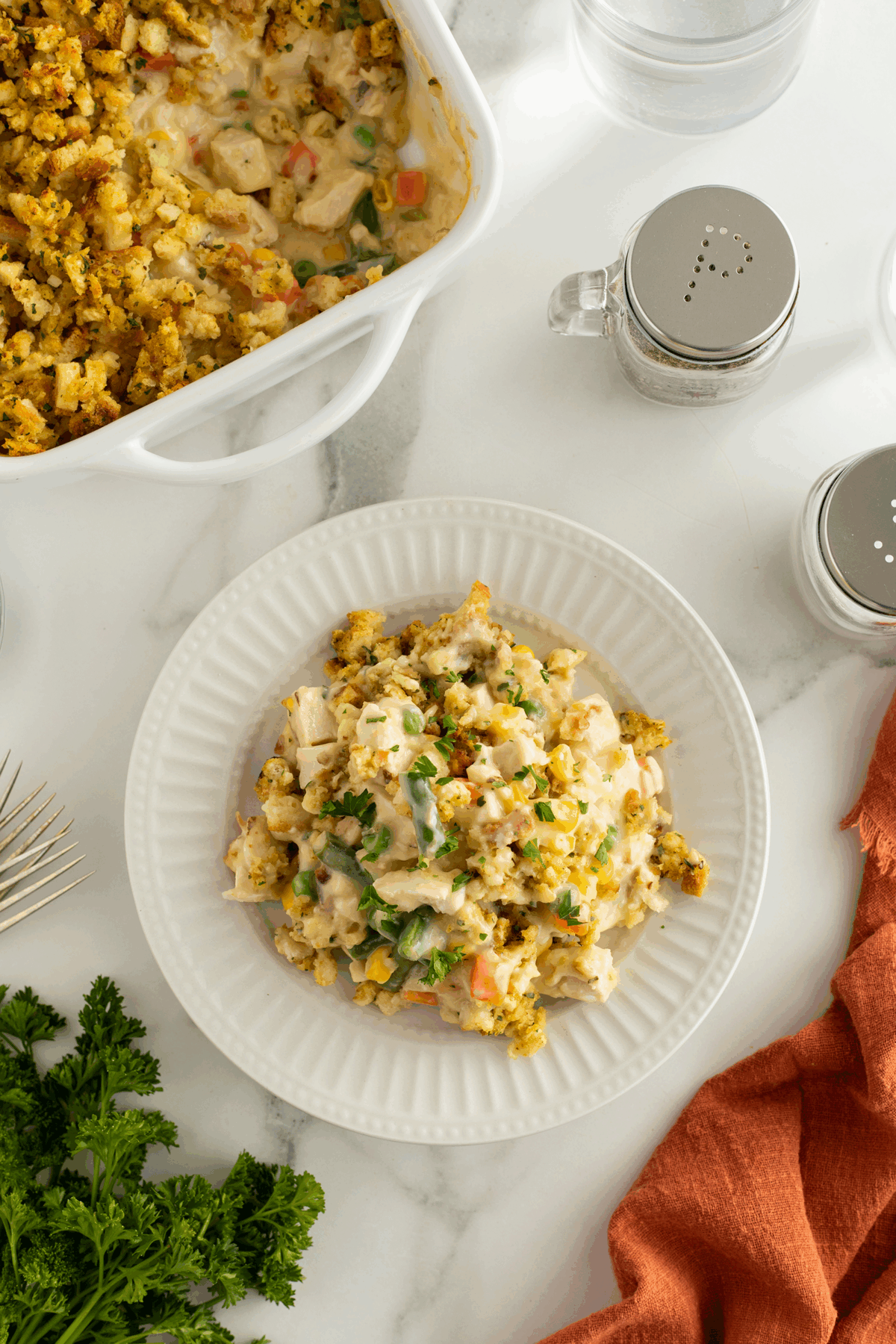 A plate of creamy chicken casserole topped with stuffing, featuring mixed vegetables, sits beside a casserole dish on a marble table with salt, pepper, a fork, orange napkin, and fresh parsley.