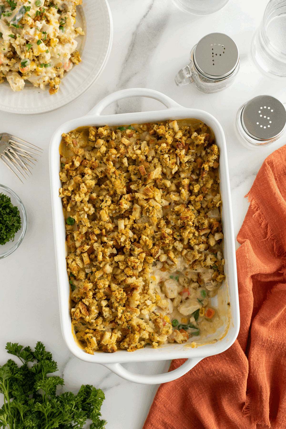 A white casserole dish filled with a baked casserole topped with golden stuffing sits on a marble counter beside an orange cloth, parsley, a plate of casserole, and salt and pepper shakers.