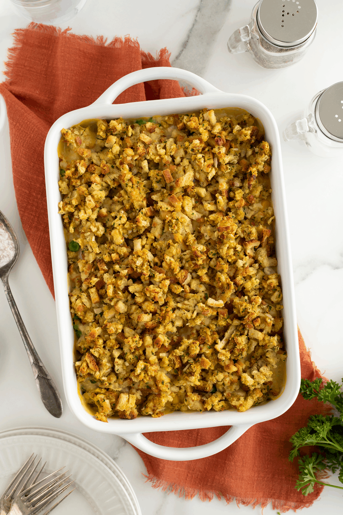A white baking dish filled with golden-brown stuffing casserole sits on an orange cloth. Nearby are a serving spoon, plates with forks, salt and pepper shakers, and a sprig of fresh parsley.