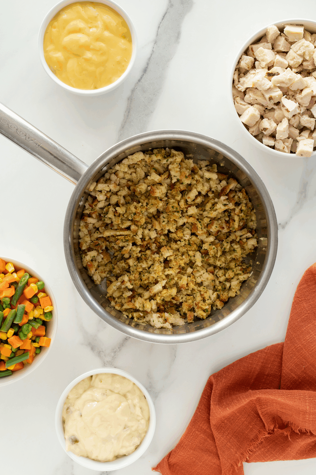 A saucepan filled with seasoned stuffing sits on a marble surface, surrounded by bowls of chopped cooked chicken, mixed vegetables, cream of chicken soup, and mayonnaise. An orange napkin is nearby.