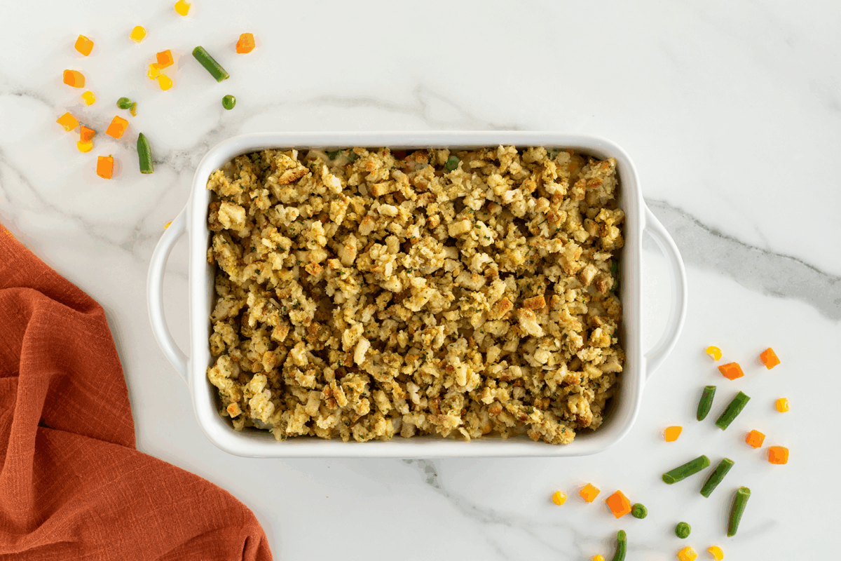 A white casserole dish filled with seasoned stuffing sits on a marble surface. Diced vegetables, including carrots, corn, and green beans, are scattered around, and an orange cloth is partially visible on the left.