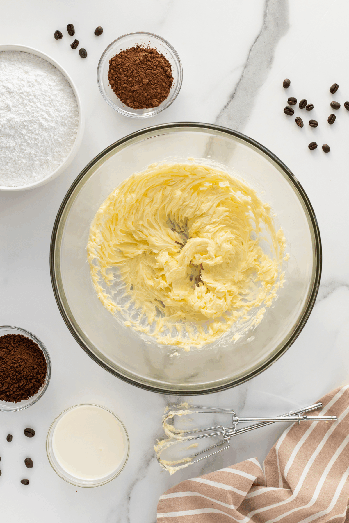 A glass bowl of creamed butter sits on a marble counter, surrounded by bowls of cocoa powder, powdered sugar, instant coffee, a glass of milk, coffee beans, a striped cloth, and a hand mixer with beaters.