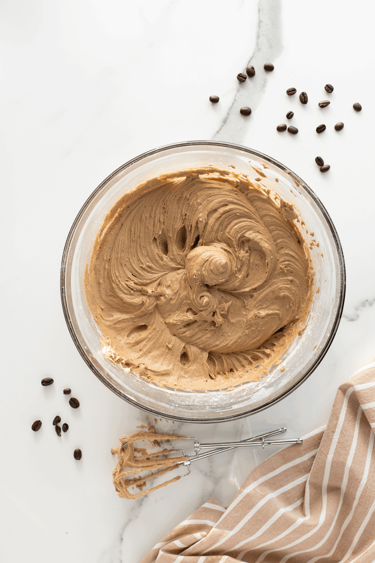A glass bowl filled with creamy brown batter, surrounded by scattered coffee beans and chocolate chips on a white marble surface. A metal whisk and a beige-striped cloth are also visible beside the bowl.