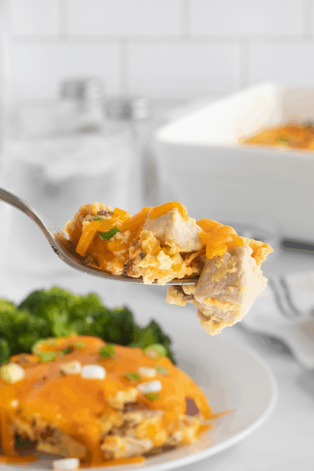 A close-up of a fork holding a bite of cheesy casserole with sausage and green onions. In the background, a plate with more casserole and broccoli is visible, along with a baking dish and salt and pepper shakers.