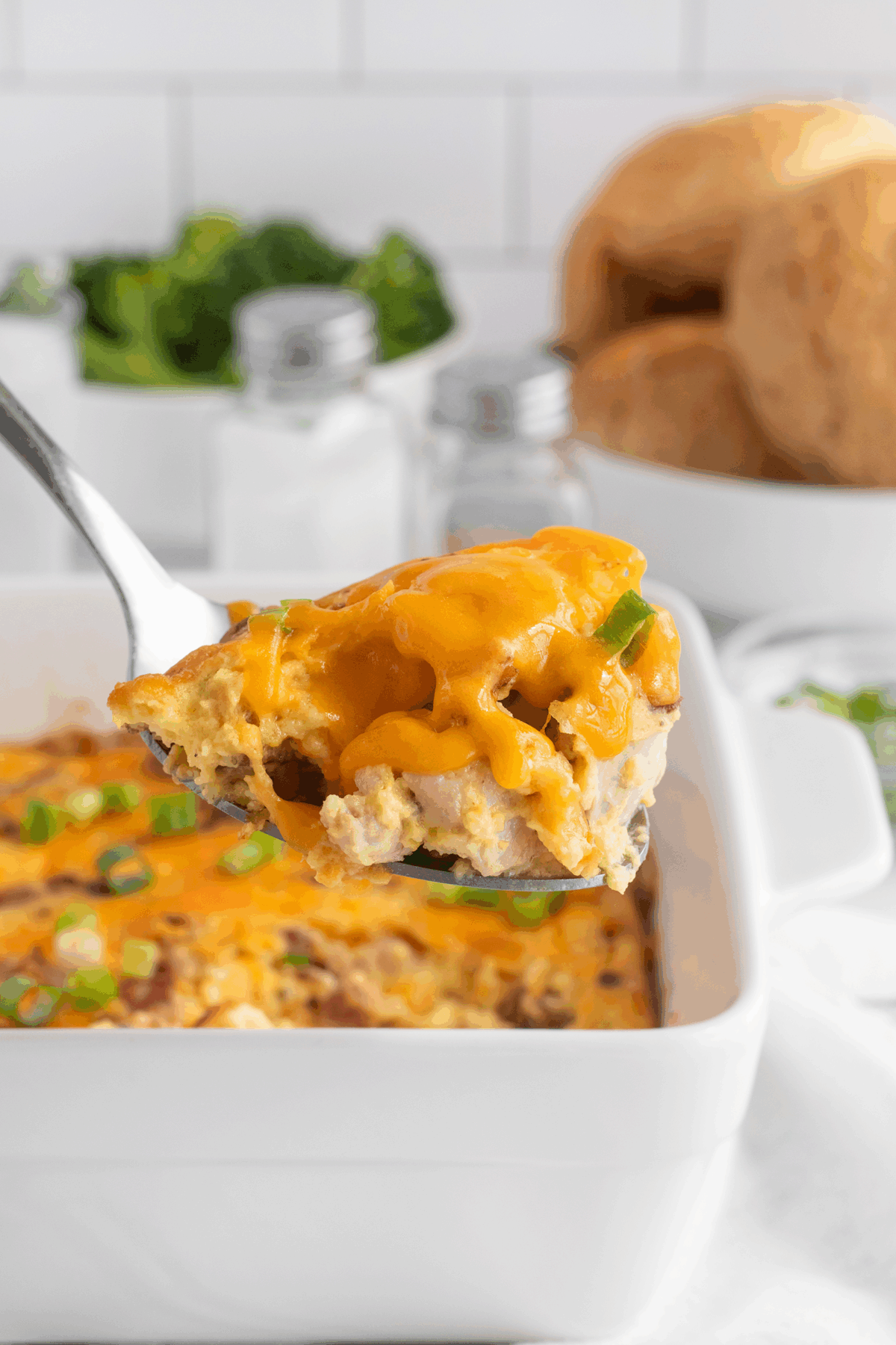A close-up of a serving spoon lifting a cheesy breakfast casserole topped with melted cheddar from a white baking dish, with bread rolls and salt and pepper shakers in the background.
