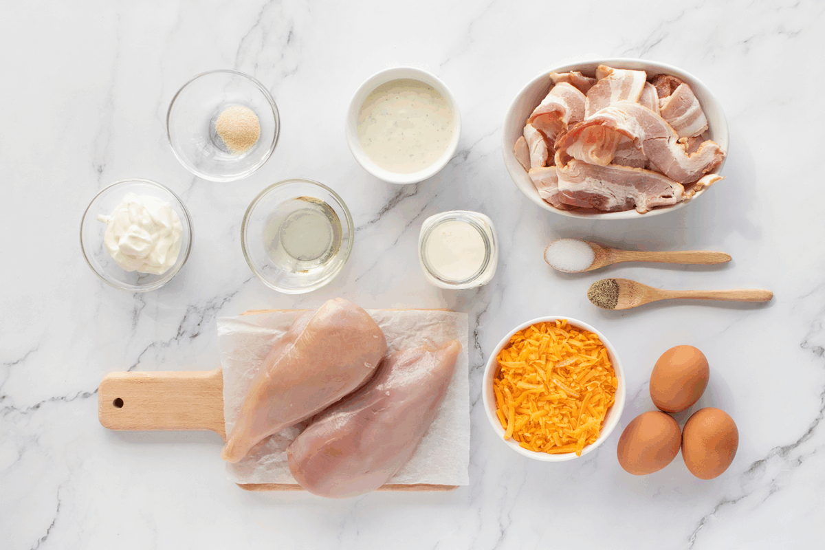Top-down view of ingredients for a recipe, including raw chicken breasts, shredded cheddar cheese, eggs, bacon, seasonings, and small bowls of mayonnaise, a creamy sauce, and oil, all arranged on a white marble surface.