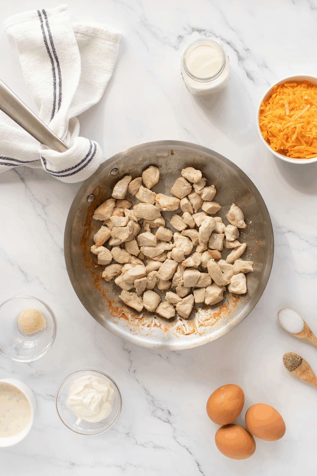 A skillet with cooked chicken pieces surrounded by ingredients in bowls: shredded cheddar cheese, eggs, sour cream, milk, seasonings, and a kitchen towel on a white marble surface.