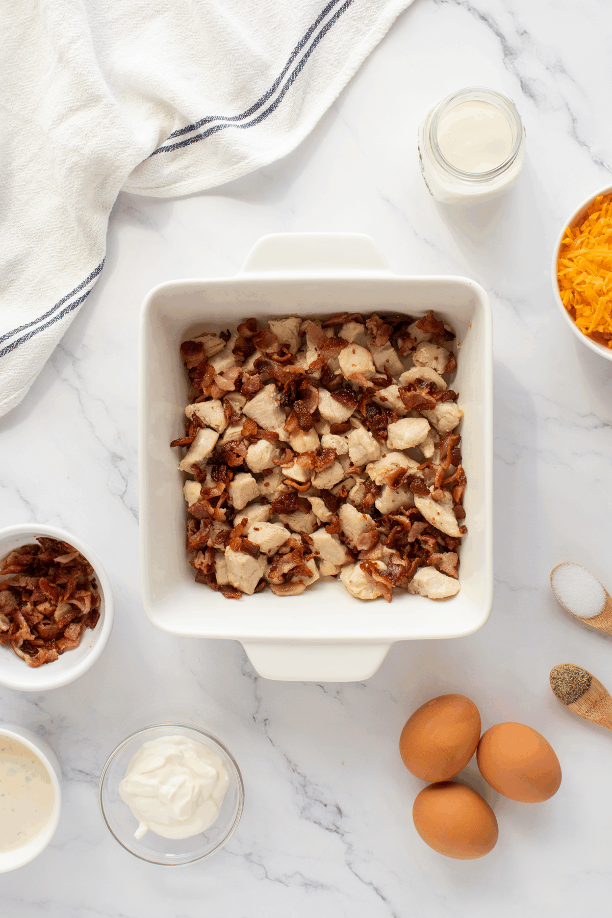 A white baking dish filled with chopped cooked chicken and bacon sits on a marble counter, surrounded by eggs, shredded cheese, sour cream, a jar of milk, seasonings, and a white towel.