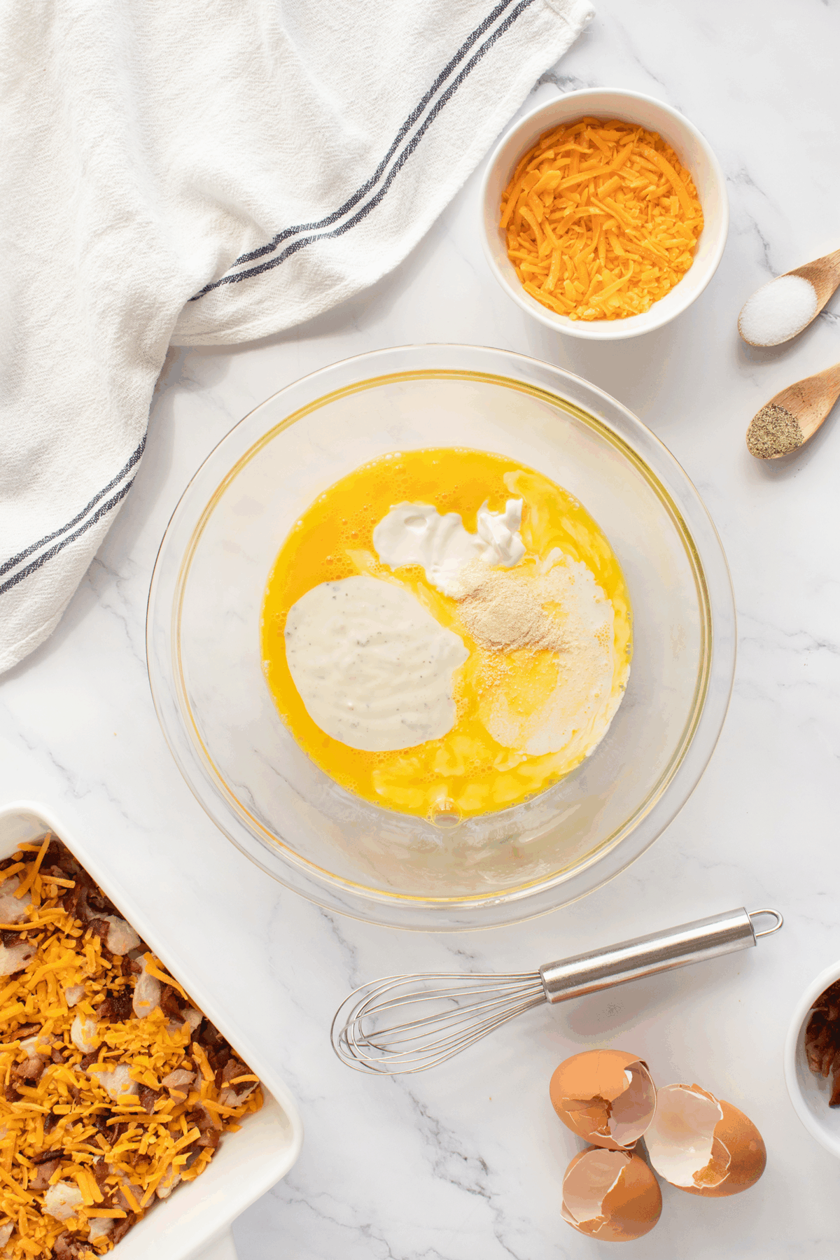 A glass bowl with melted butter, sour cream, ranch seasoning, and cheese sits on a marble countertop. Nearby are shredded cheese, eggshells, a whisk, a towel, and a casserole dish with cheese and bacon.