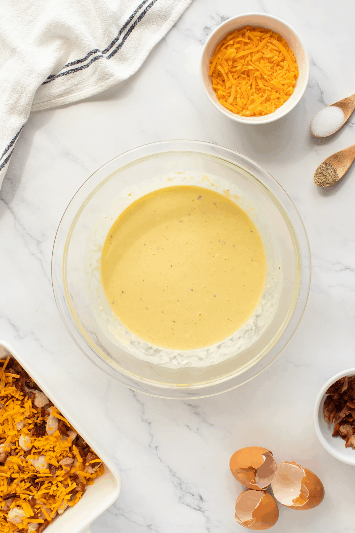 A glass bowl of creamy egg mixture sits on a marble countertop, surrounded by bowls of shredded cheddar cheese, salt, pepper, crumbled bacon, and onion, with cracked eggshells nearby.