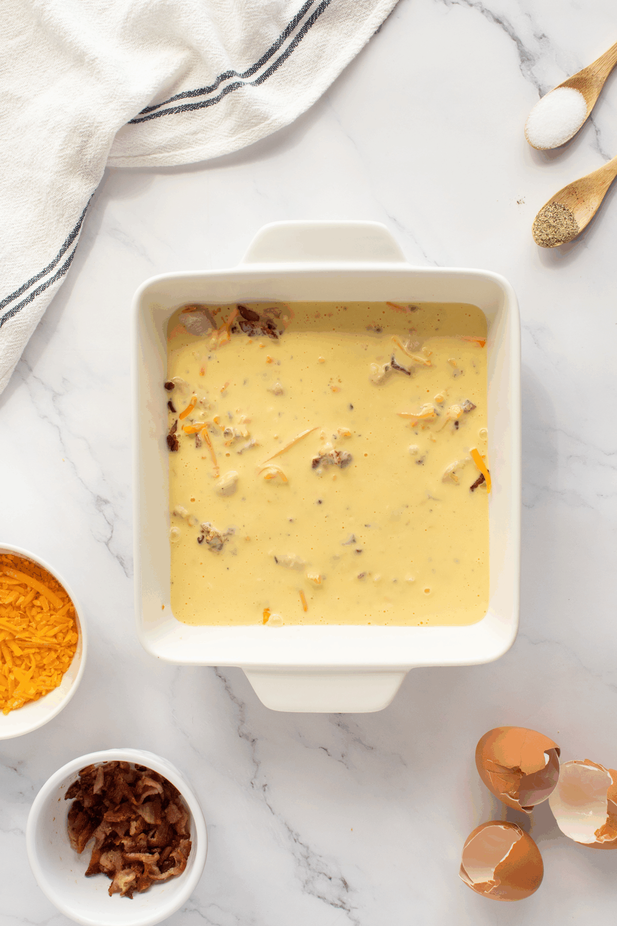 A square white baking dish filled with an egg and cheese mixture sits on a marble countertop, surrounded by shredded cheddar, crumbled bacon, eggshells, and a striped kitchen towel.
