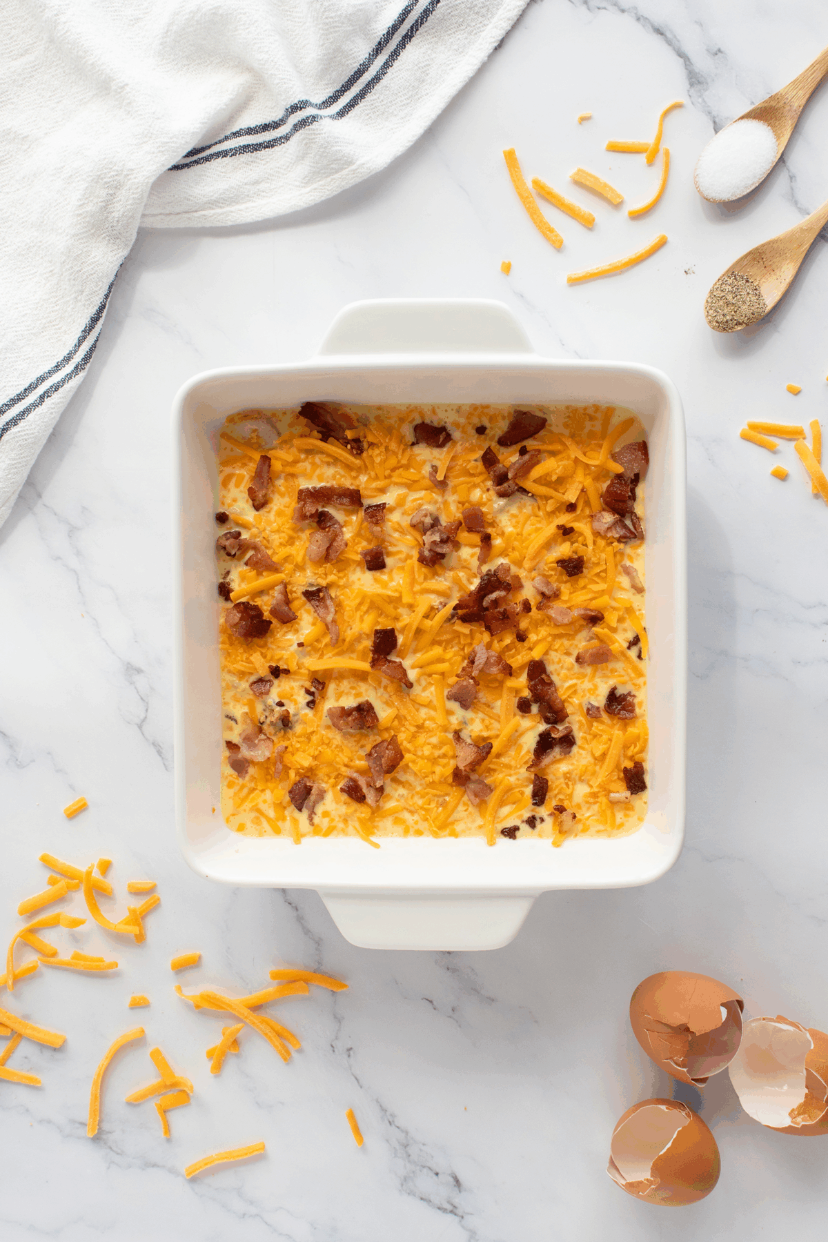 A square white baking dish filled with a mixture of shredded cheddar cheese and bacon bits on a marble countertop, surrounded by scattered cheese, eggshells, seasonings, and a white kitchen towel.
