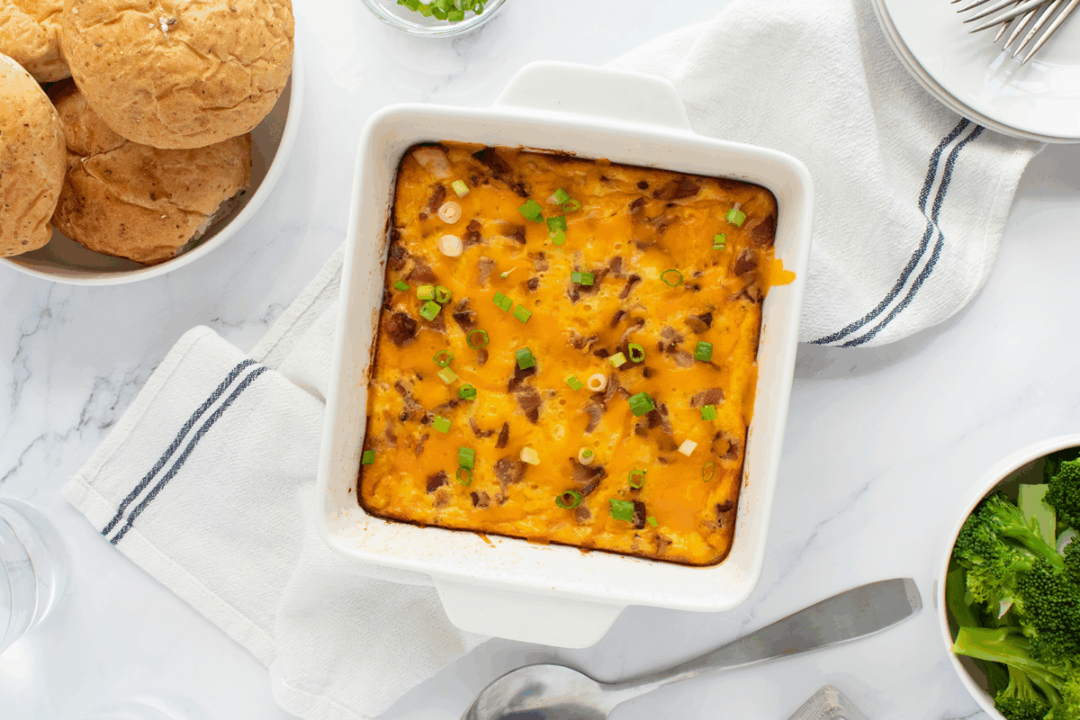 A baked casserole topped with melted cheddar cheese, green onions, and bacon bits sits in a white dish. Surrounding it are rolls, plates, utensils, and a bowl of broccoli on a white table with a striped towel.