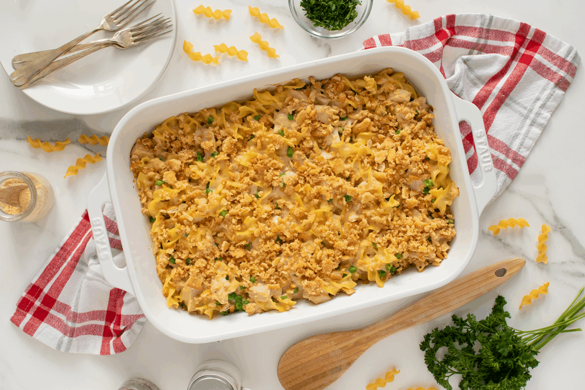 A white casserole dish filled with a baked noodle casserole topped with crushed crackers sits on a marble surface, surrounded by parsley, a red and white towel, plates, forks, and uncooked spiral pasta.