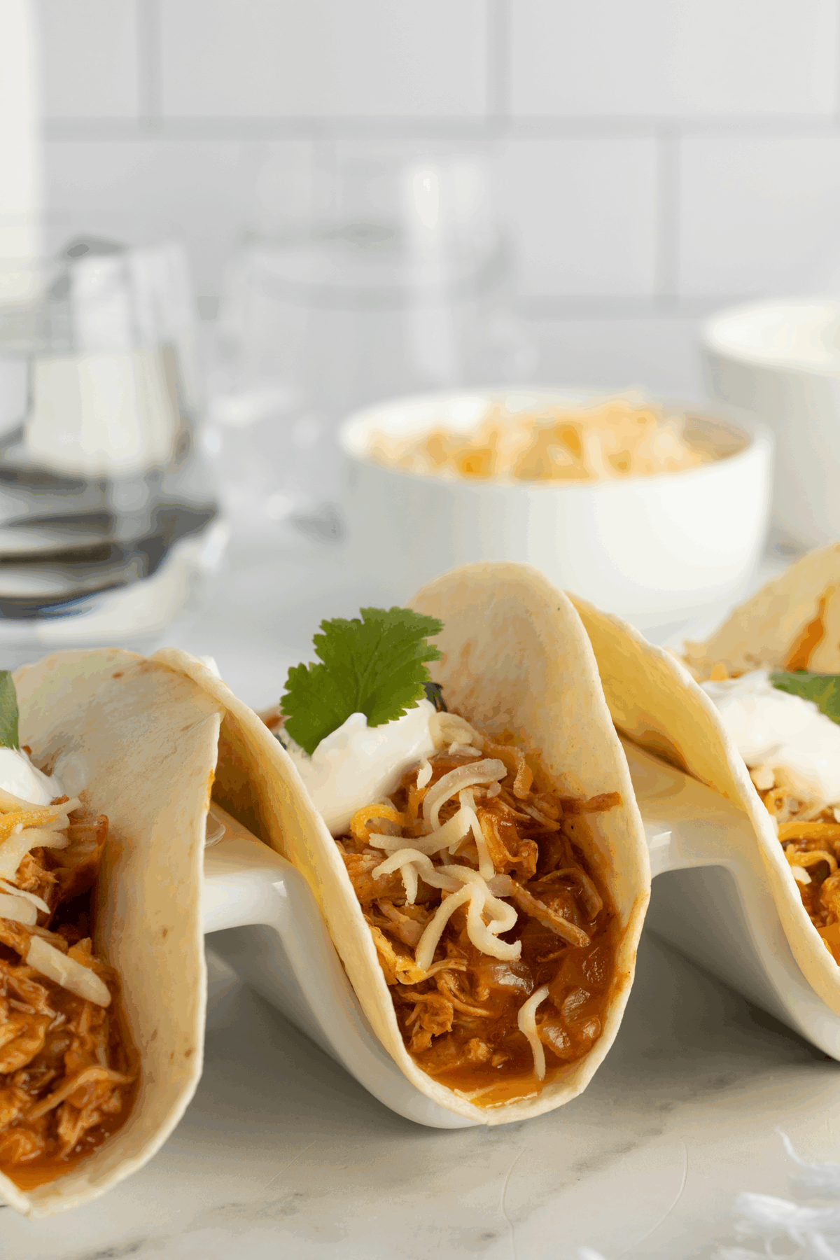 Three soft tacos filled with shredded chicken, melted cheese, and topped with sour cream and cilantro are displayed in a white taco holder. Blurred bowls and glasses are in the background on a white surface.