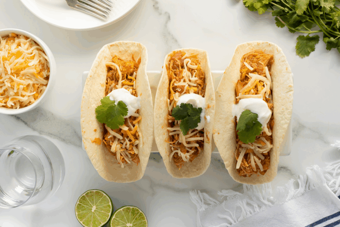 Three taco boats filled with shredded chicken, topped with shredded cheese, sour cream, and cilantro are arranged on a white tray. Lime halves, shredded cheese, and fresh cilantro are nearby on a marble surface.