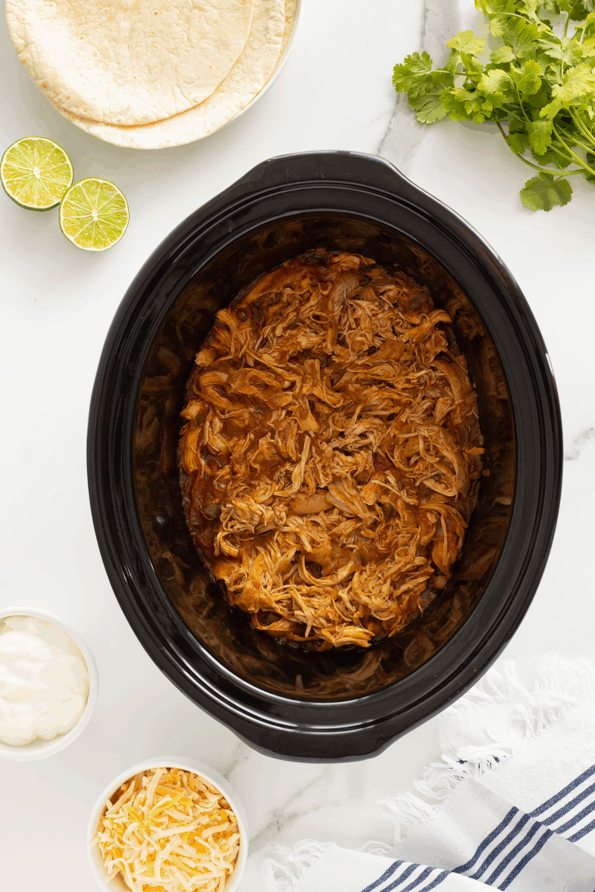 Shredded chicken in a black slow cooker, surrounded by tortillas, lime halves, fresh cilantro, a bowl of shredded cheese, a bowl of sour cream, and a blue-striped towel on a white surface.