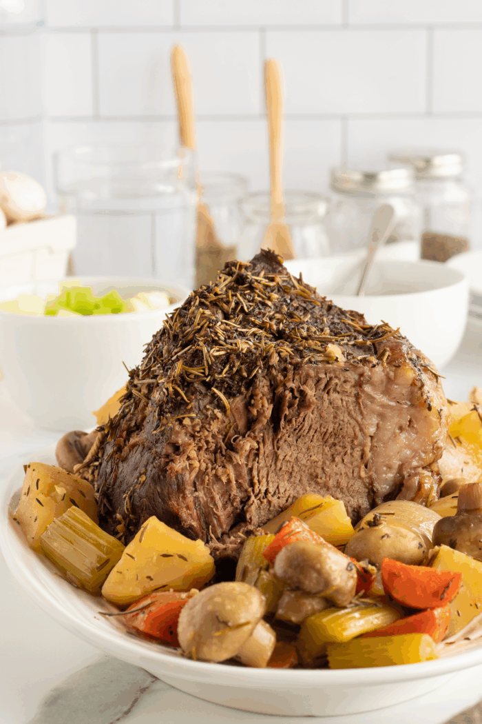 A cooked pot roast garnished with herbs is served on a white plate, surrounded by carrots, potatoes, celery, and mushrooms, with kitchen jars and utensils in the background.