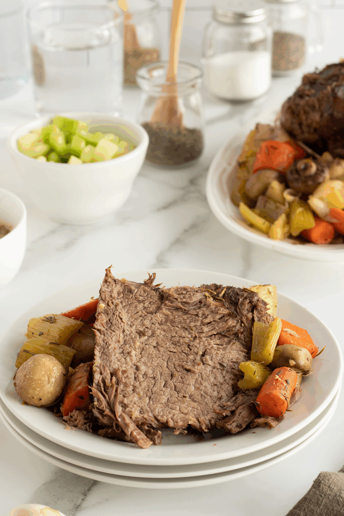 A plate of pot roast with shredded beef, potatoes, carrots, celery, and mushrooms sits on a white table. In the background, there are bowls of chopped celery and jars of seasonings.