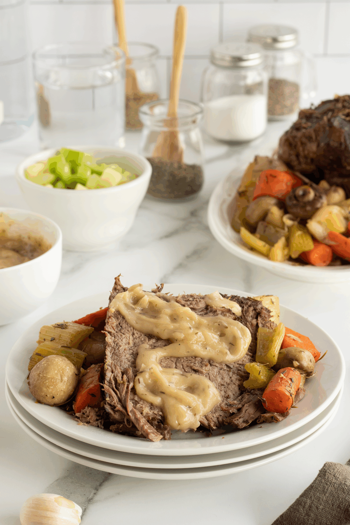 A plate with sliced pot roast topped with gravy, surrounded by cooked carrots, potatoes, and celery. In the background are bowls of diced celery, sauce, seasonings, and two glasses of water on a marble countertop.