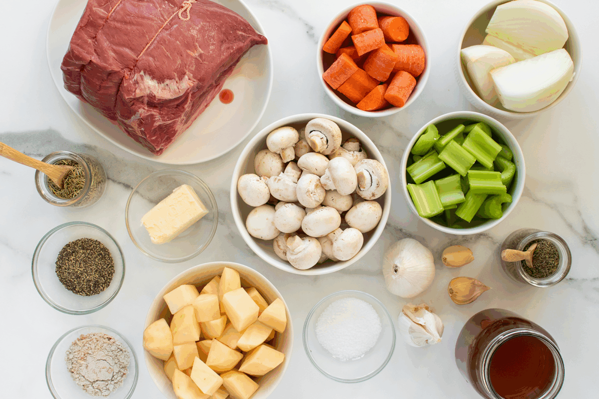 A top-down view of raw pot roast ingredients: a slab of beef, chopped carrots, potatoes, onions, celery, whole mushrooms, garlic, butter, herbs, flour, salt, and broth arranged in bowls on a white surface.