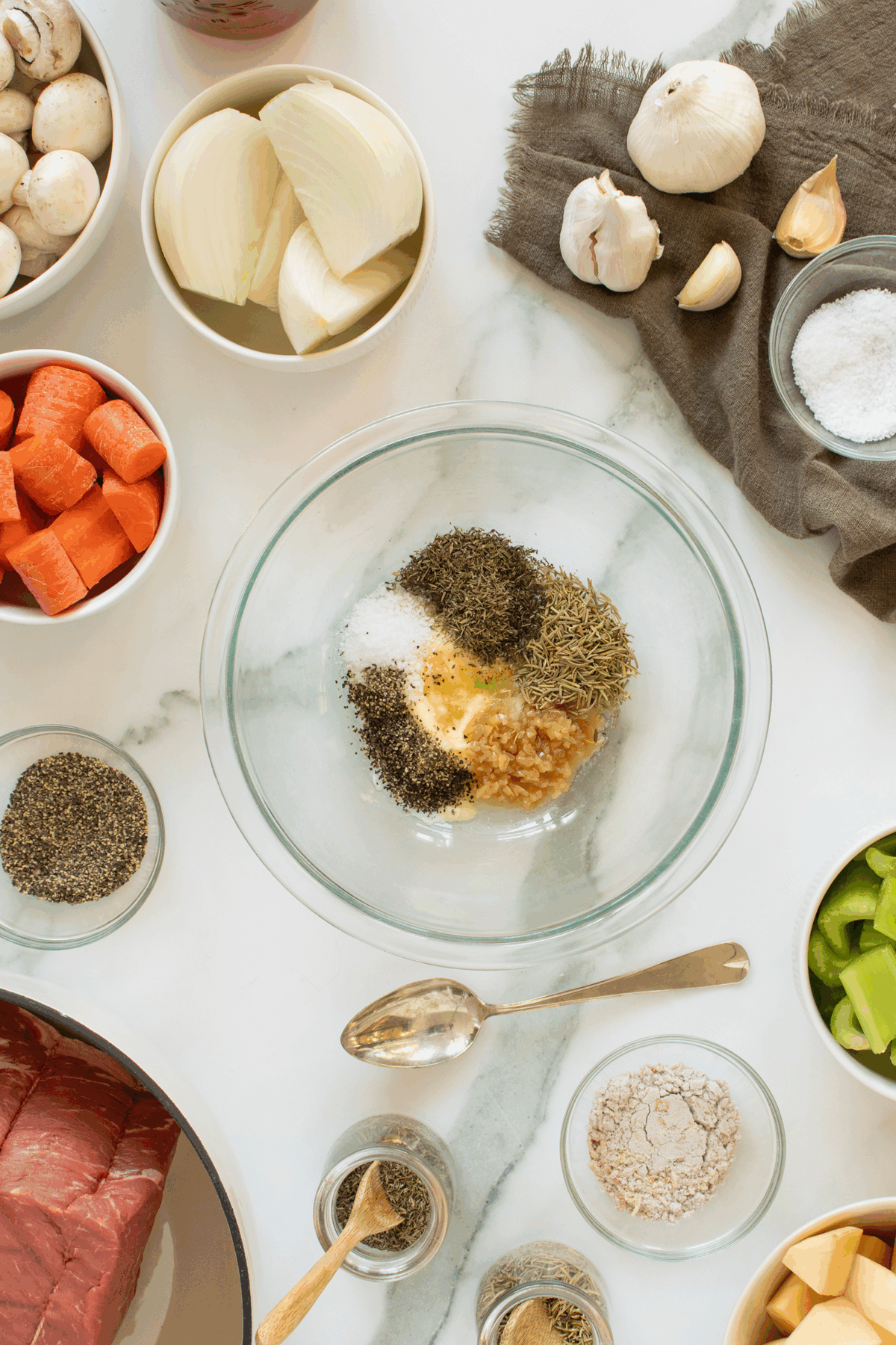 A glass bowl with dried herbs, salt, pepper, and minced garlic sits on a marble counter surrounded by raw vegetables, a piece of beef, and various seasonings in small bowls.