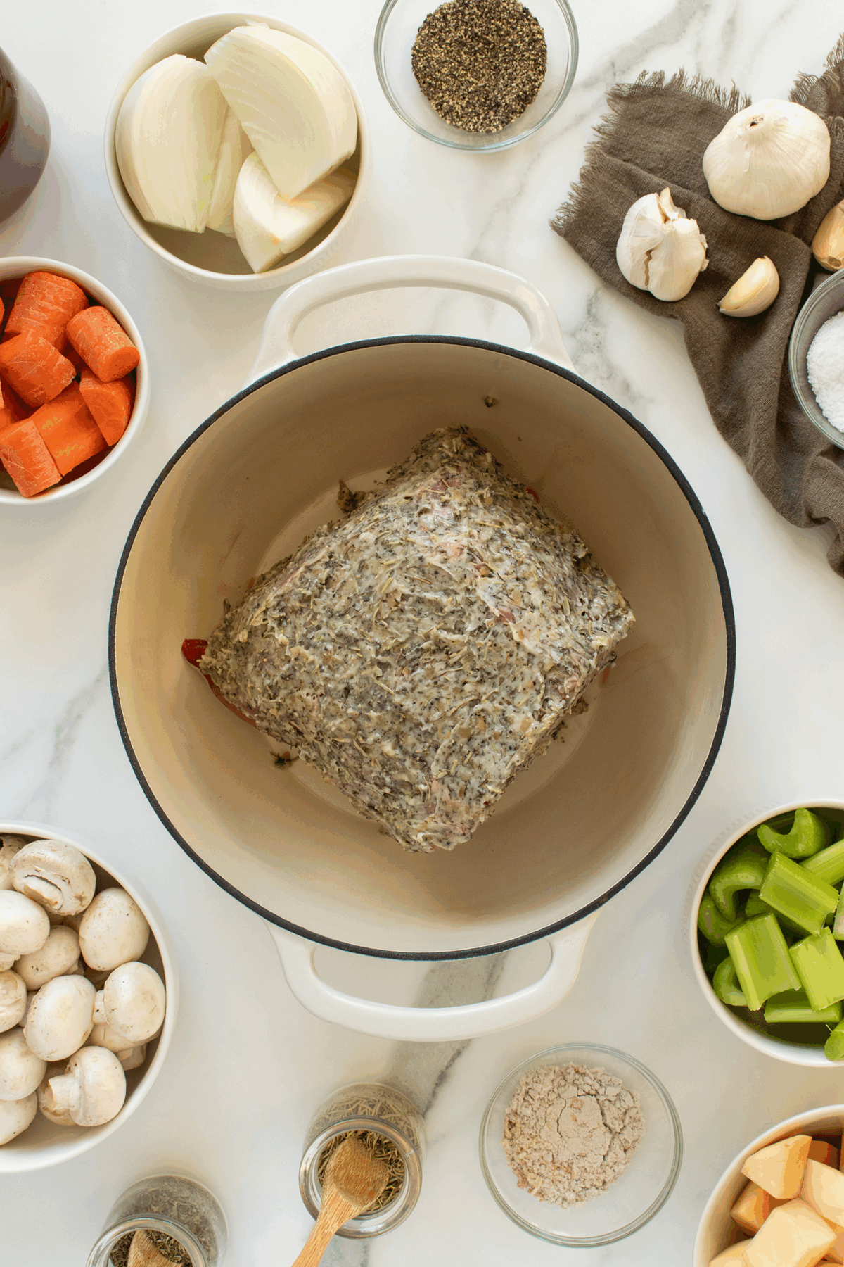 Overhead view of a pot with a seasoned meat roast surrounded by bowls of chopped onions, carrots, celery, mushrooms, garlic, spices, and a towel on a marble countertop.