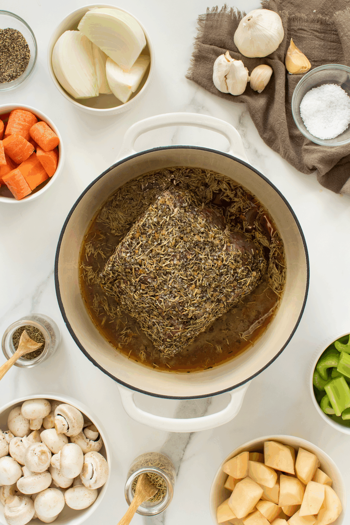 Overhead view of a herb-crusted beef roast cooking in a Dutch oven surrounded by bowls of raw mushrooms, carrots, celery, rutabaga, onion, salt, garlic, black pepper, and seasonings on a white surface.