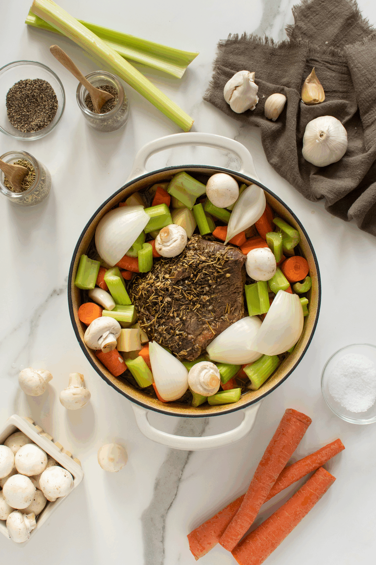 A pot with a beef roast, carrots, celery, onions, and mushrooms sits on a white surface surrounded by garlic, black pepper, herbs, salt, mushrooms, carrots, and a dark cloth napkin.