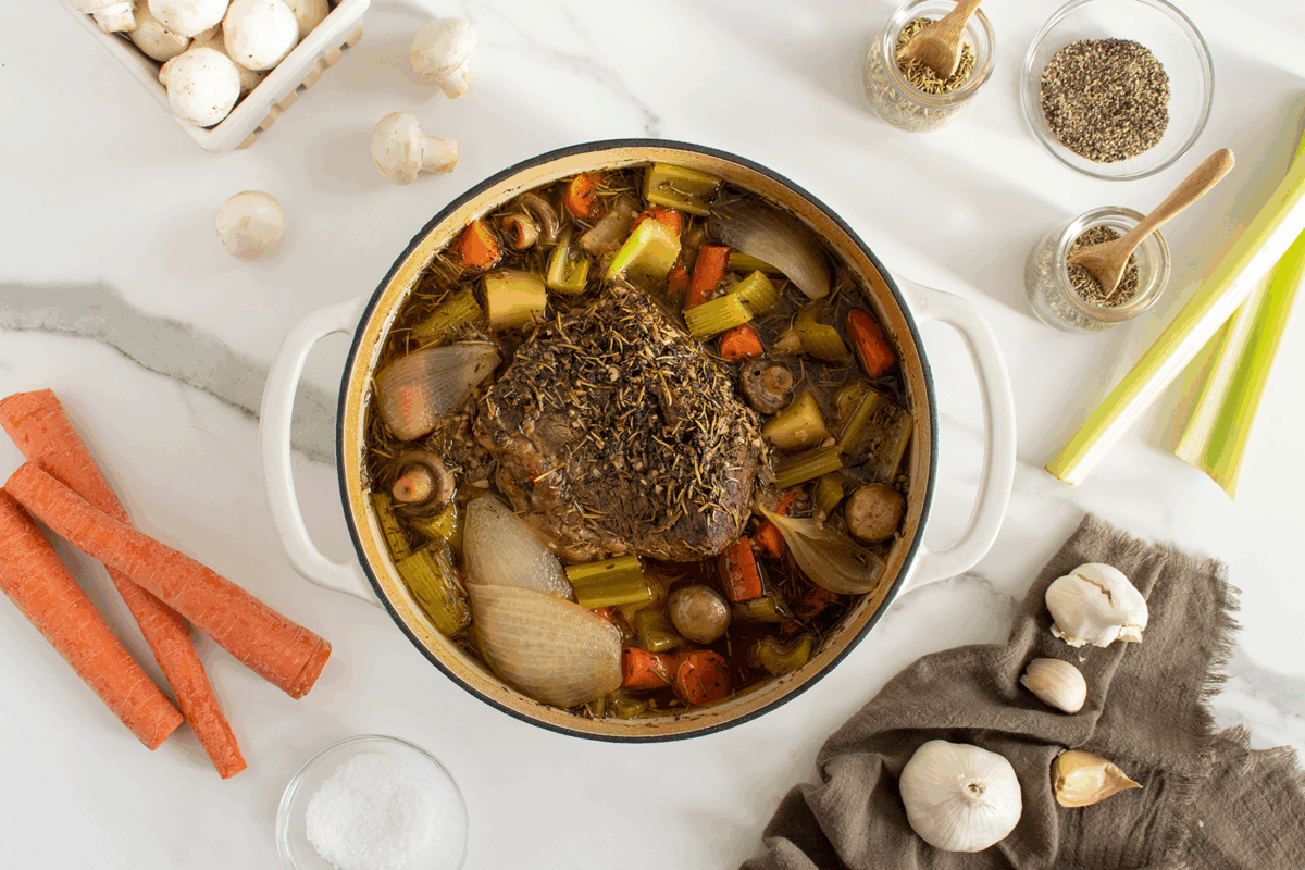 A pot roast with herbs, carrots, celery, onions, and mushrooms in a white Dutch oven, surrounded by raw vegetables, seasonings, and a salt bowl on a marble countertop.