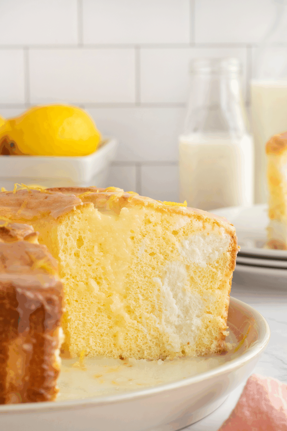 A sliced lemon-flavored cake with a creamy filling sits on a plate. In the background, lemons, a glass bottle of milk, and a glass of milk are visible on a white kitchen counter with subway tile backsplash.