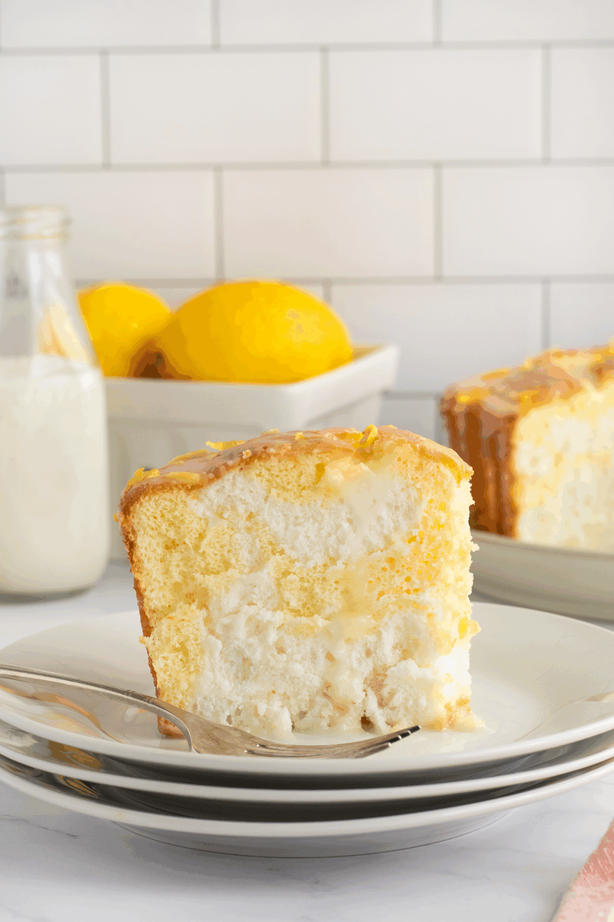 A slice of lemon cake with glaze sits on a plate with a fork. In the background, there’s a bottle of milk and a bowl of whole lemons, with part of the remaining cake visible. The scene has a bright, clean kitchen feel.