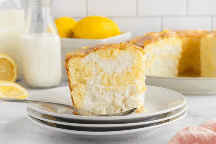 A slice of lemon angel food cake with creamy filling on stacked white plates, a fork beside it. In the background are fresh lemons, a bottle of milk, and the remaining cake on a platter.