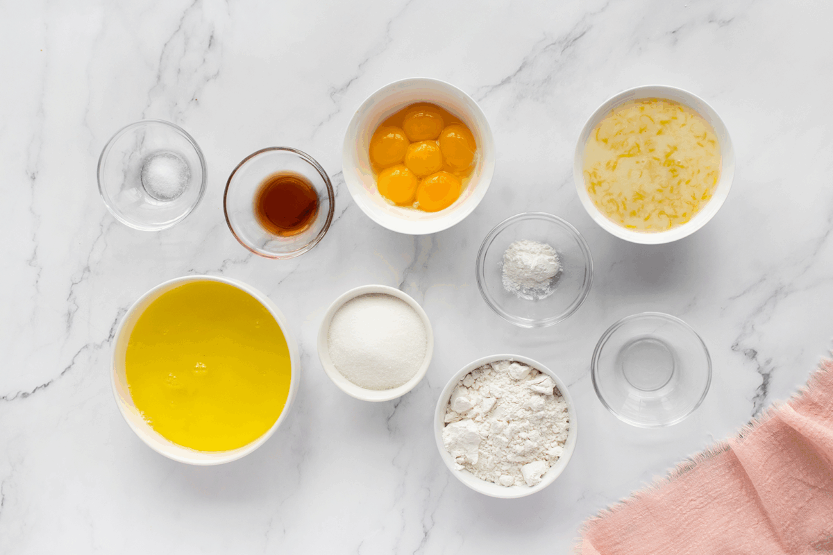 Top-down view of bowls with baking ingredients on a marble surface: egg yolks, egg whites, flour, sugar, milk with lemon zest, vanilla extract, salt, cream of tartar, and a pink cloth in the corner.