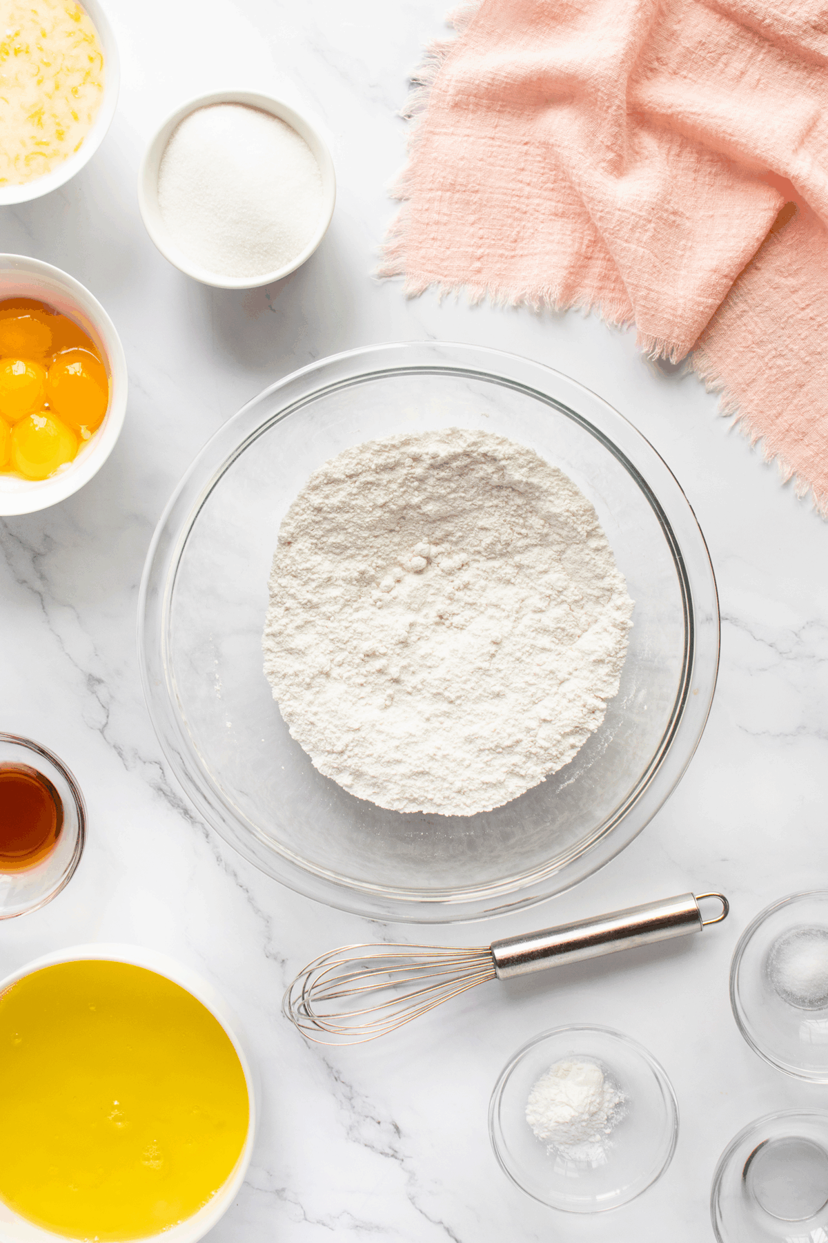 A glass bowl filled with flour sits on a marble surface, surrounded by bowls of eggs, sugar, melted butter, vanilla, baking powder, and a whisk. A pink cloth lies in the corner.