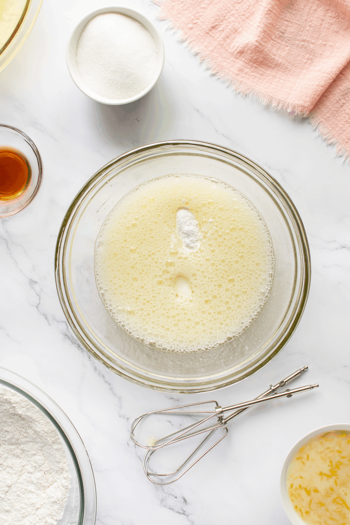 A glass bowl with a frothy mixture sits on a marble countertop, surrounded by bowls of sugar, flour, melted butter, vanilla, and a metal whisk, next to a pink cloth.