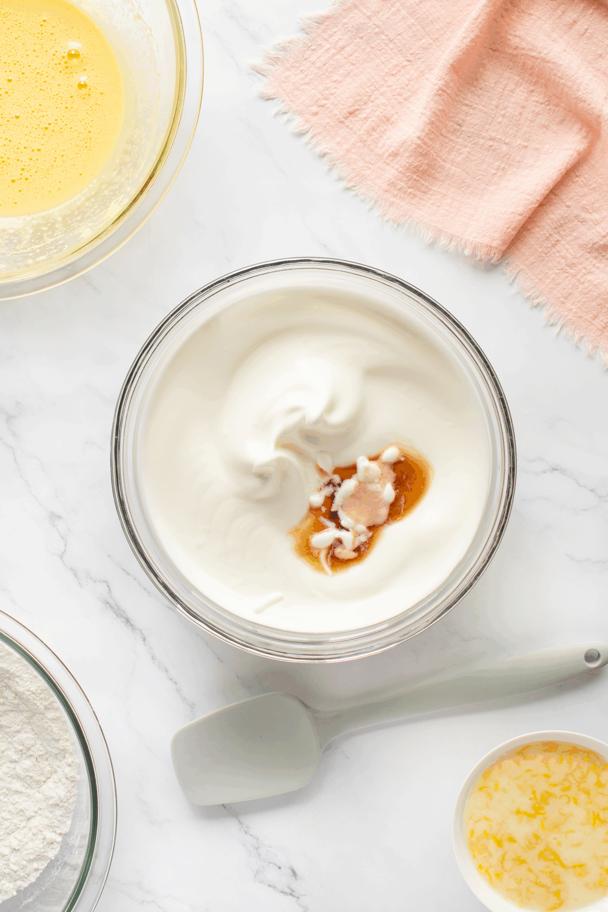 A glass bowl filled with whipped cream and a dollop of vanilla extract, surrounded by bowls of beaten eggs, flour, melted butter, and a spatula on a marble countertop with a pink cloth.