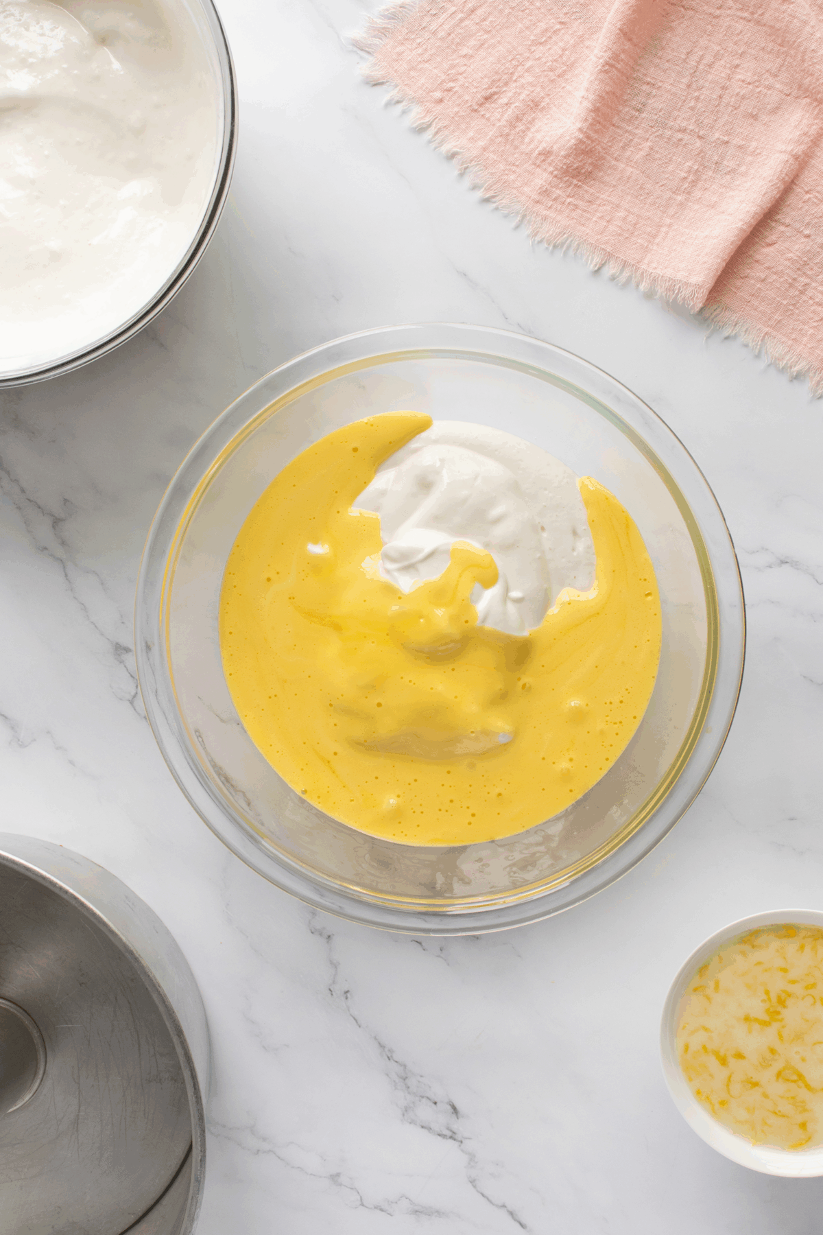 A glass bowl with a yellow mixture and a dollop of white cream sits on a marble counter. Nearby are a metal bowl with more cream, a small bowl with grated lemon zest, and a pink cloth.