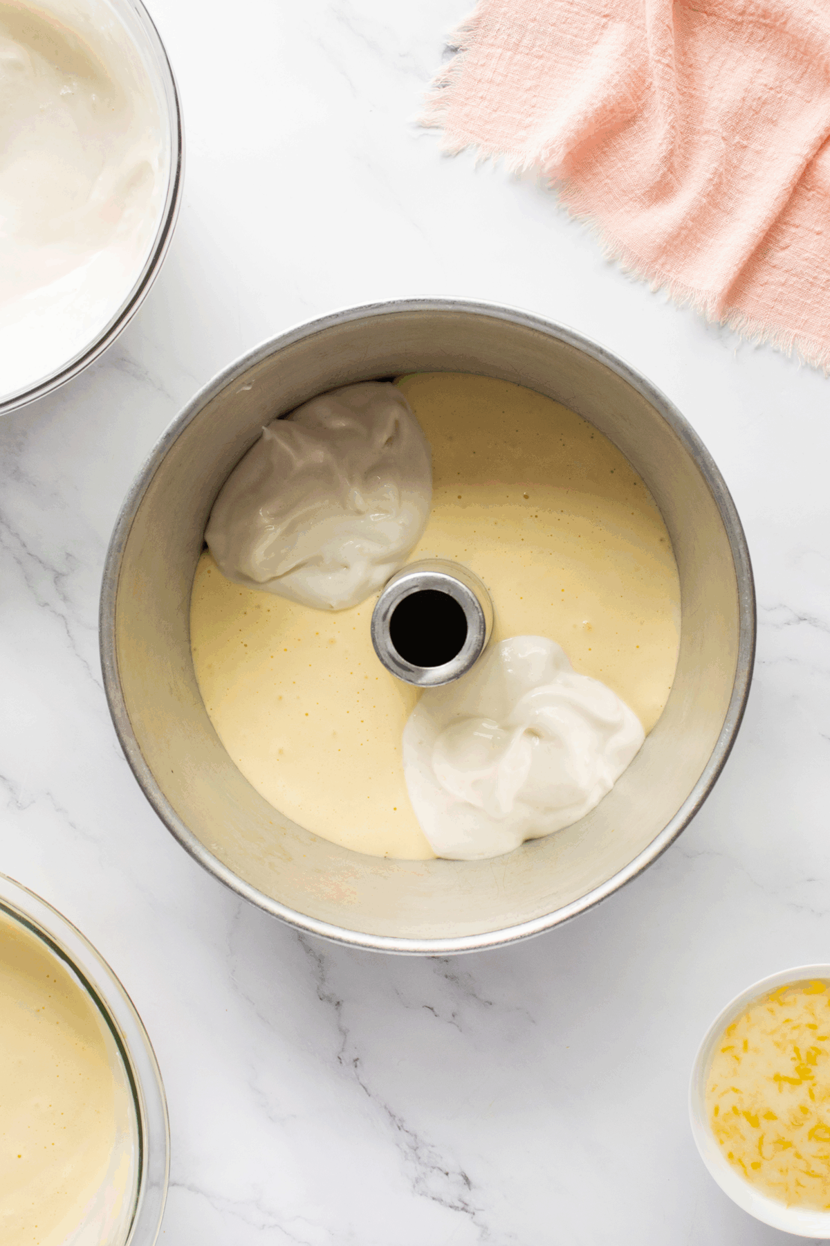 A bundt pan filled with yellow cake batter and two dollops of white whipped mixture, surrounded by bowls and a pink cloth on a marble surface.