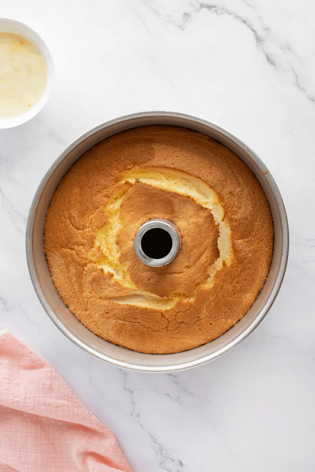A golden-brown pound cake baked in a round bundt pan sits on a white marble surface. A bowl with white liquid and a pink cloth are nearby.
