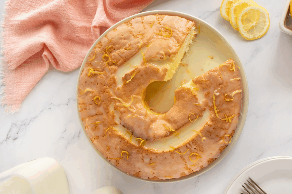 A glazed lemon bundt cake with a slice missing sits on a white plate. Lemon zest is sprinkled on top, and lemon slices, a pink cloth, and a glass of milk are nearby on a white marble surface.