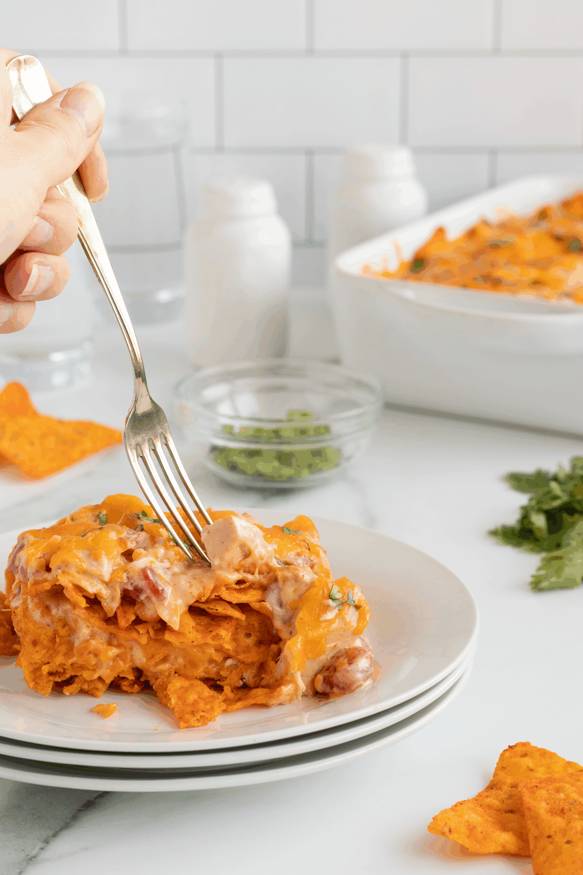 A hand holds a fork, lifting a bite from a cheesy, layered Doritos casserole on a white plate. The background shows a casserole dish, glass of water, herbs, and small bowls on a white countertop.