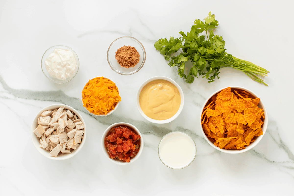 A top-down view of ingredients on a white surface, including sour cream, shredded cheese, taco seasoning, nacho cheese, cooked chicken cubes, diced tomatoes, milk, tortilla chips, and fresh cilantro.