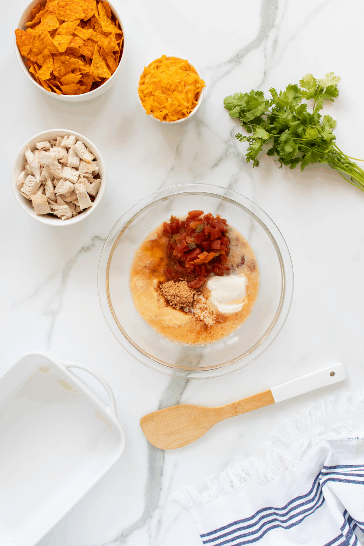 A glass bowl with salsa, spices, sour cream, and soup mix sits on a marble countertop, surrounded by bowls of shredded cheese, Doritos, cooked chicken, fresh cilantro, a spatula, and a baking dish.