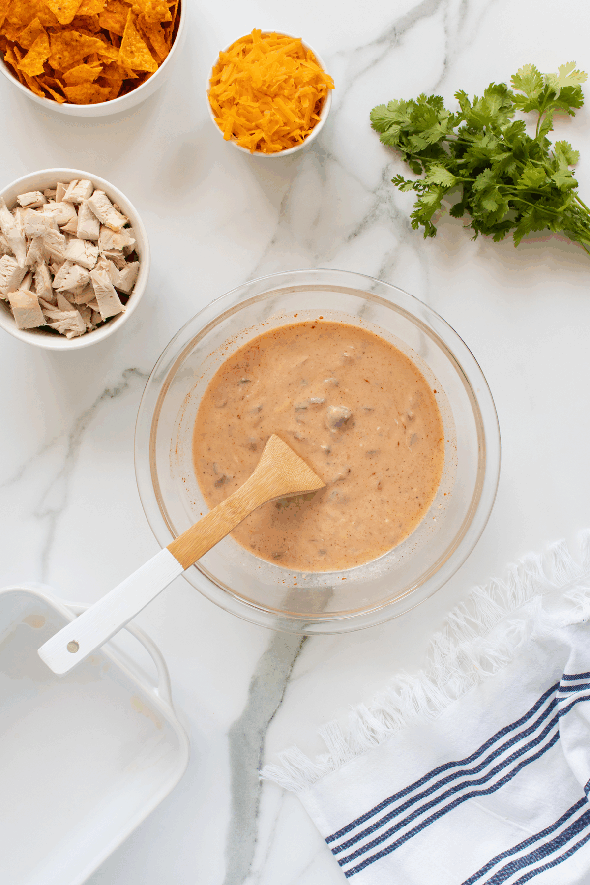A glass bowl with a creamy mixture and wooden spoon sits on a marble counter, surrounded by bowls of cubed chicken, shredded cheese, crumbled chips, fresh cilantro, and a striped towel.