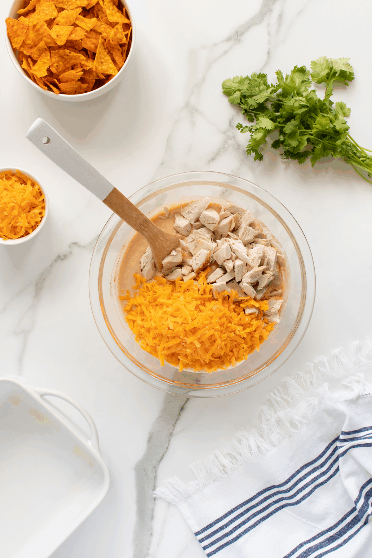 A glass bowl with chopped cooked chicken and shredded cheddar cheese on a marble counter. Nearby are bowls of crushed chips and more cheese, a bunch of cilantro, a casserole dish, and a striped towel.