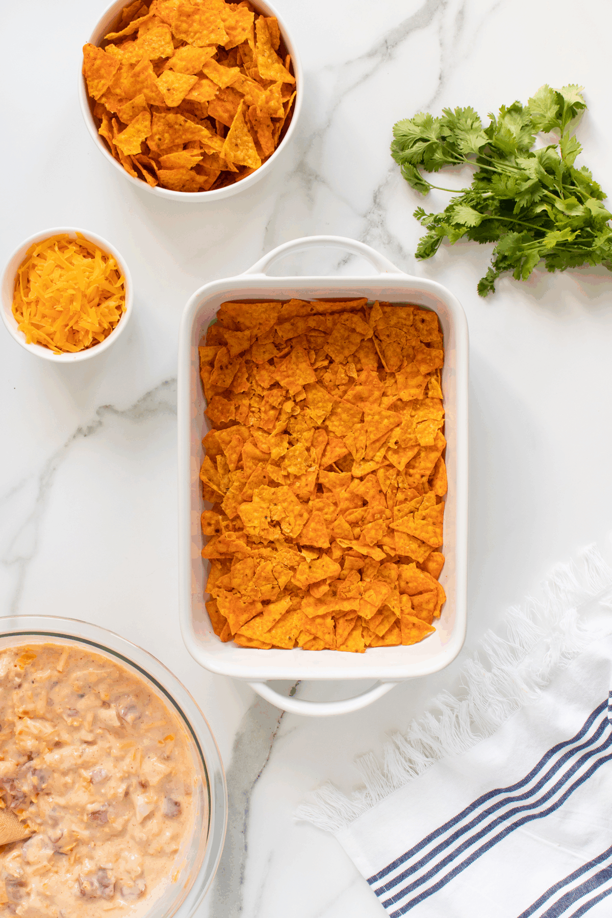 A white dish filled with crushed orange tortilla chips, surrounded by a bowl of more chips, a small bowl of shredded cheese, a glass bowl of a creamy mixture, fresh cilantro, and a striped towel on a marble surface.