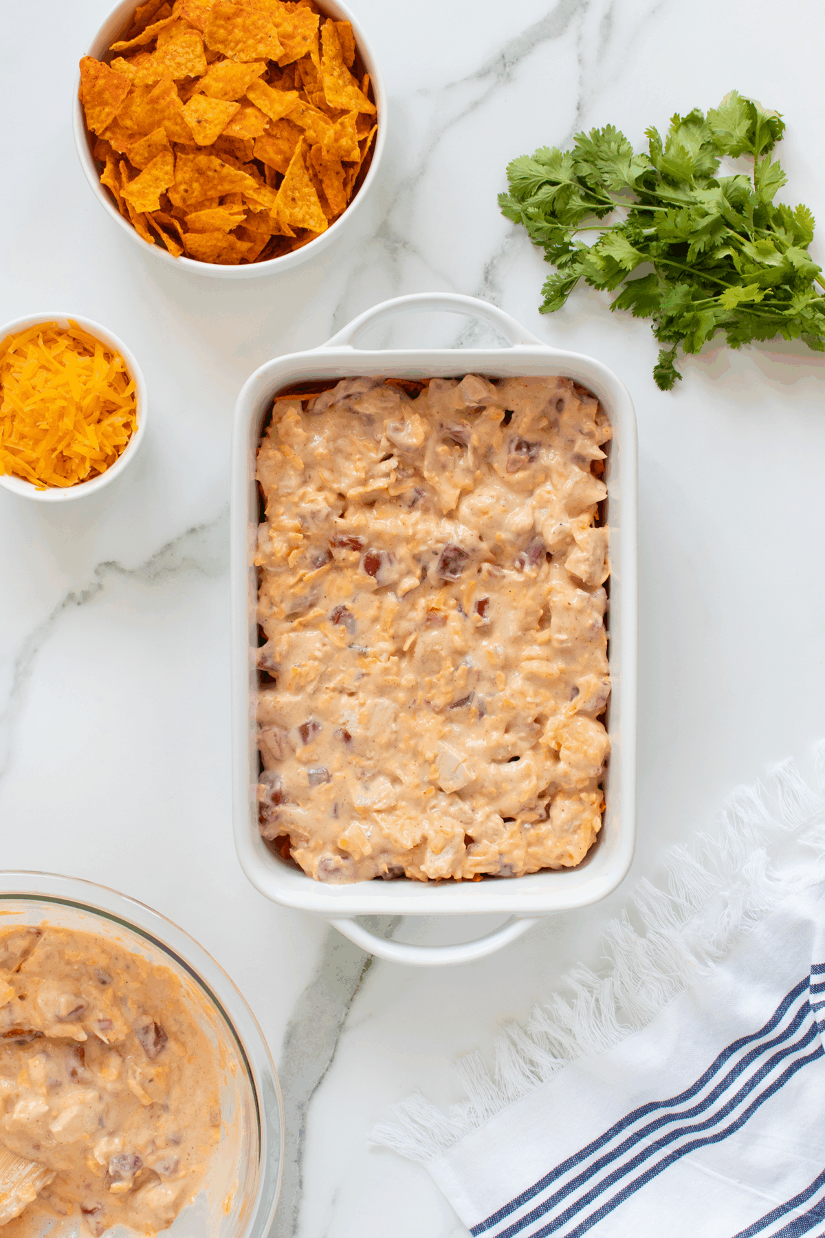 A casserole dish filled with a creamy mixture sits on a white counter, surrounded by a bowl of crushed chips, shredded cheese, chopped cilantro, and a striped towel.