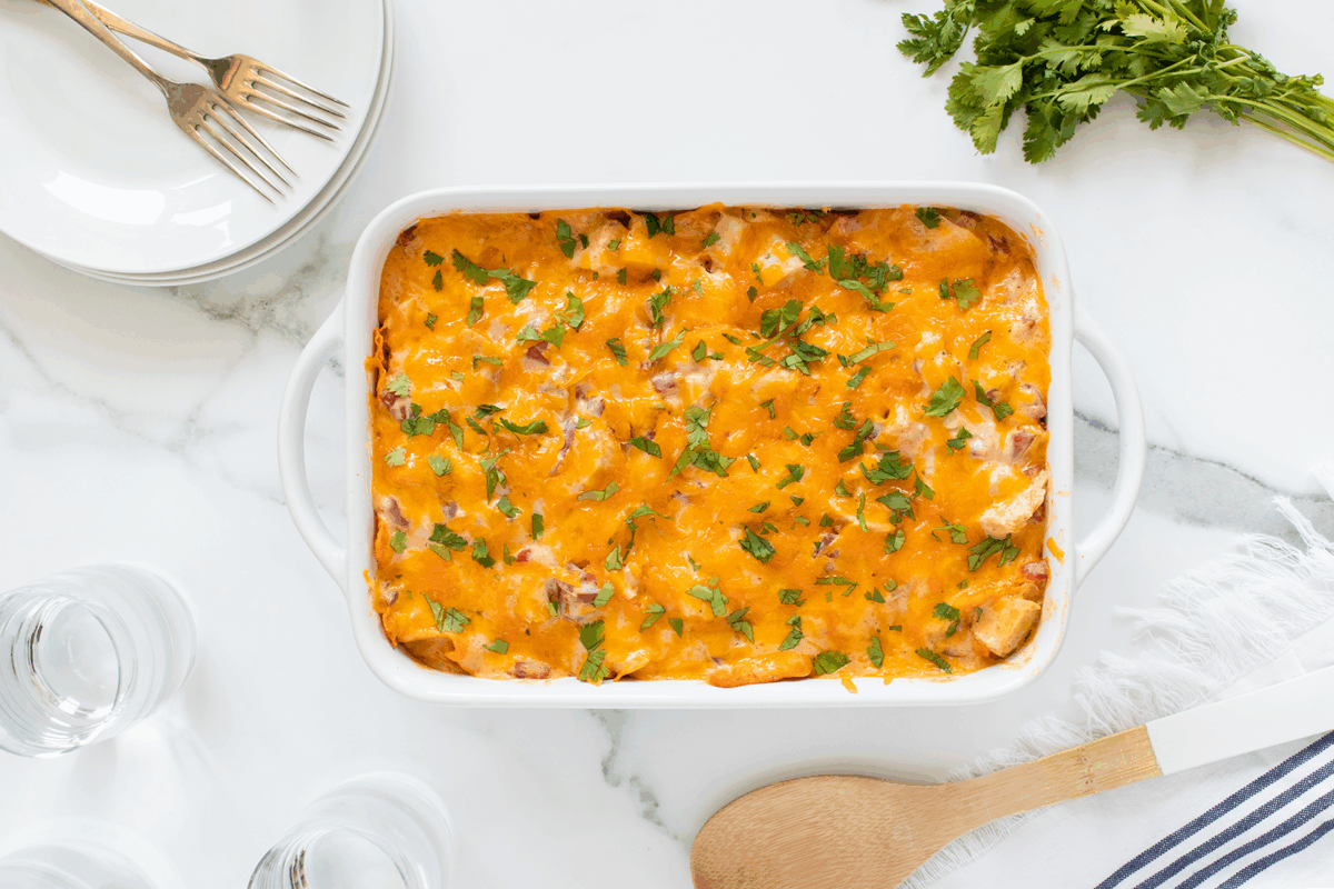 A baked casserole topped with melted cheddar cheese and chopped herbs sits in a white dish on a marble counter, near plates with forks, a bunch of parsley, and a wooden spoon.