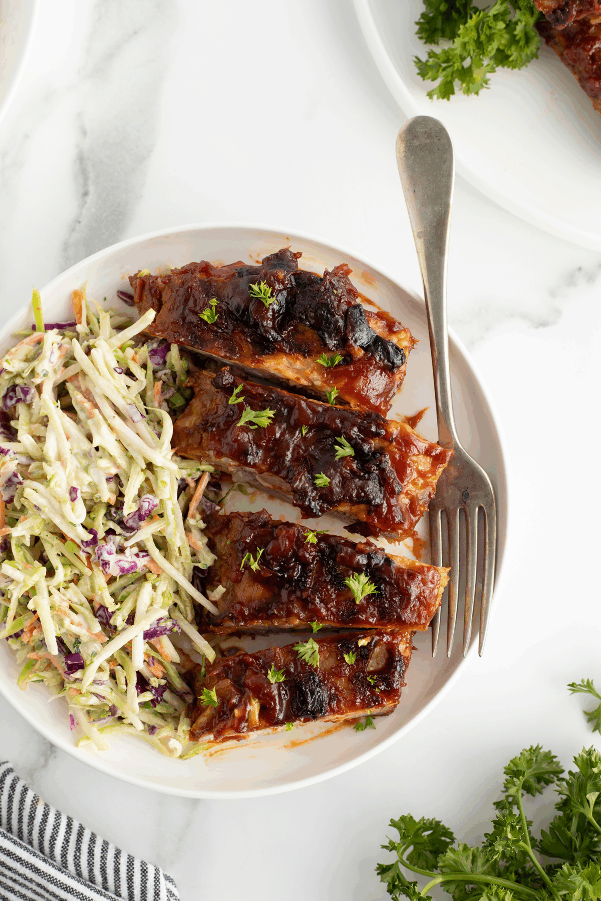 A plate with four glazed barbecue ribs topped with herbs, served alongside creamy coleslaw with shredded cabbage and carrots; a fork rests on the plate. Parsley garnish and a striped napkin are nearby.